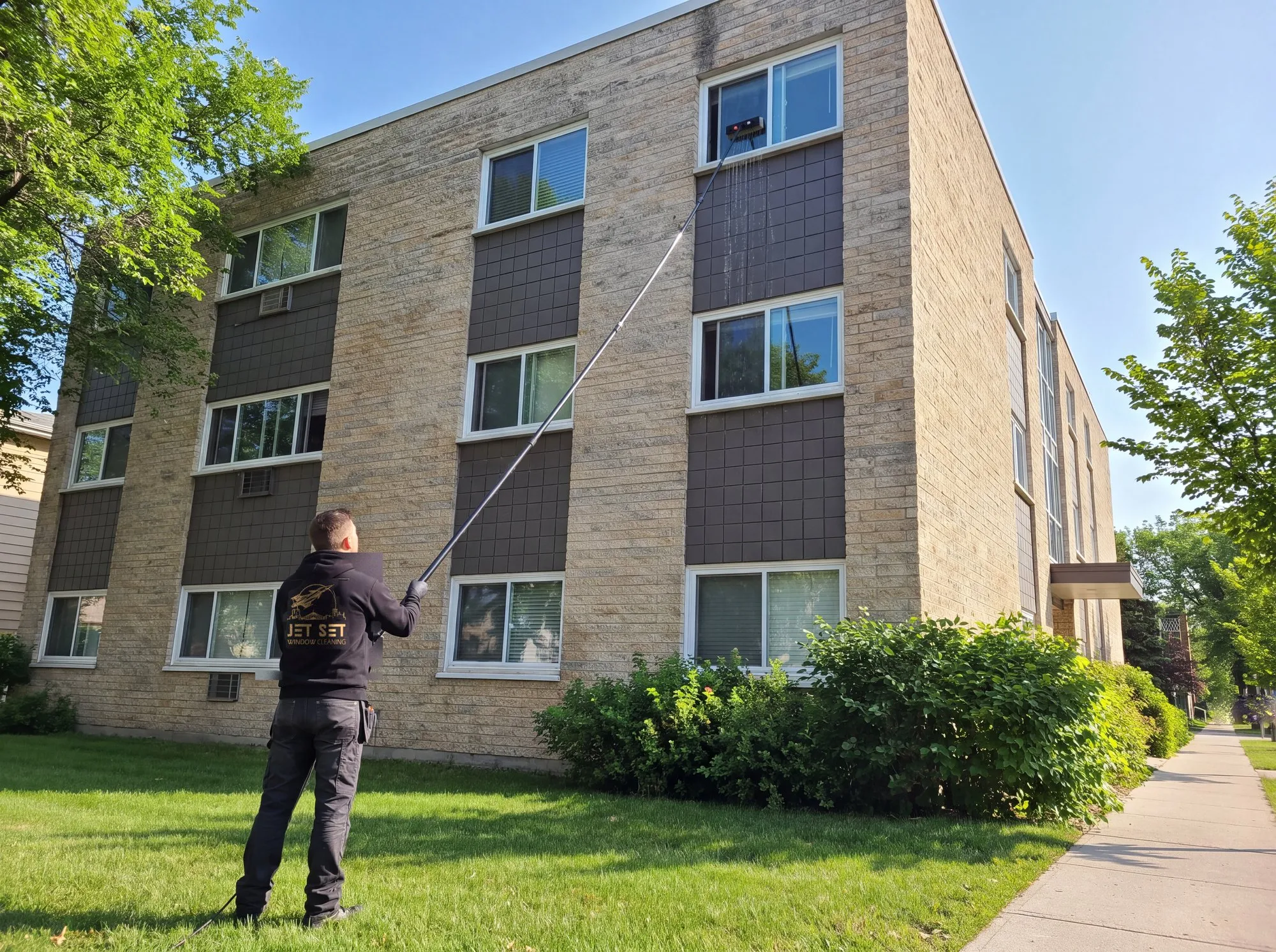 Jet Set technician on the lawn cleaning the third-floor windows of a Winnipeg apartment block with a water-fed pole