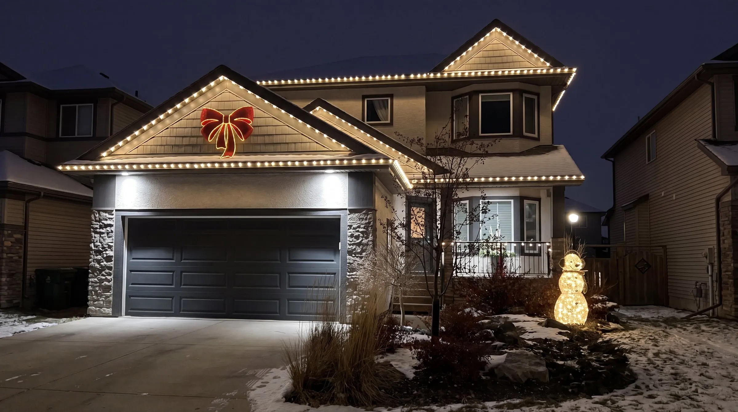 Winnipeg stucco two-storey home at night with warm white Govee permanent lights along every roofline and a lit red bow on the gable above the garage