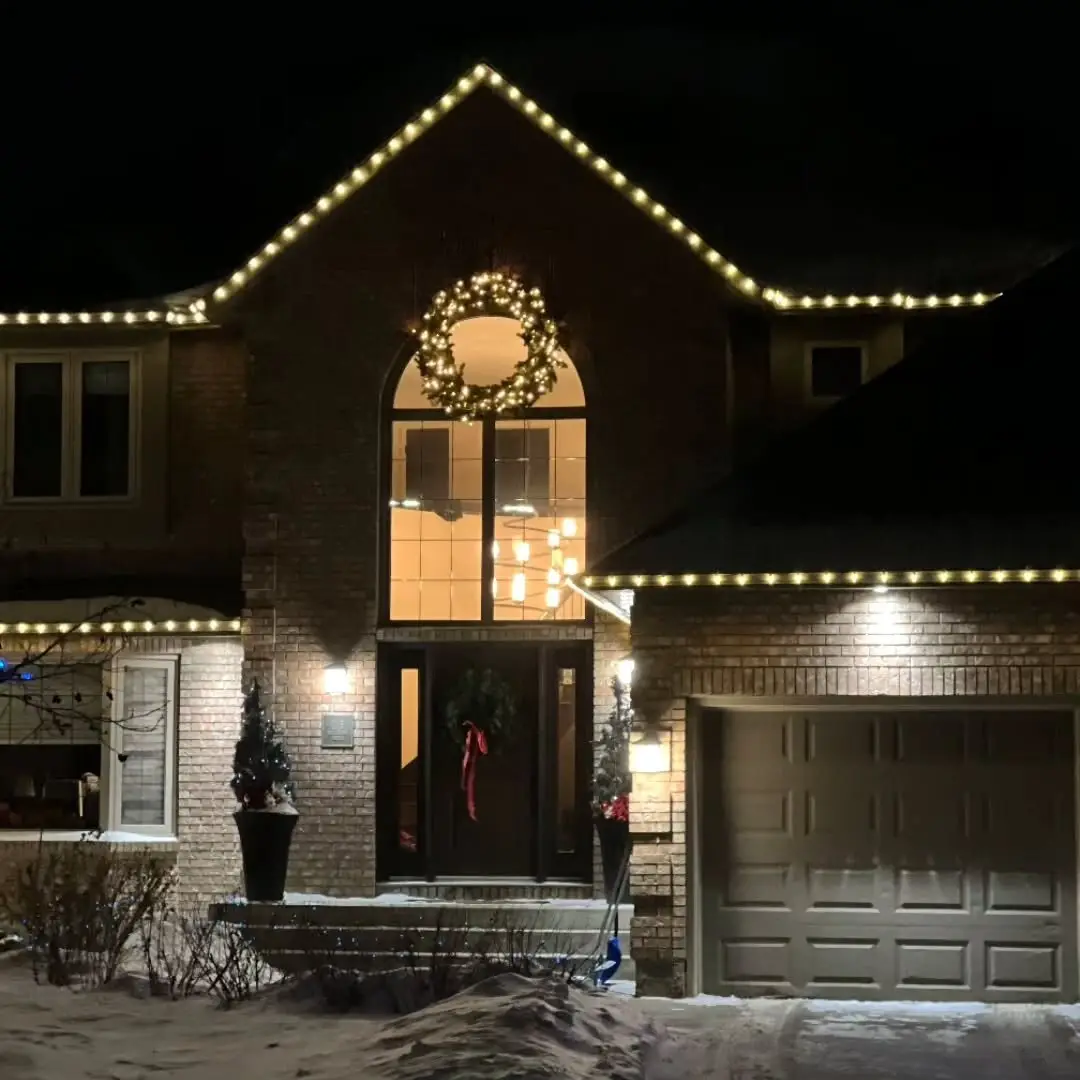 Brick two-storey home at night with warm white Christmas lights along the roofline and a lit wreath