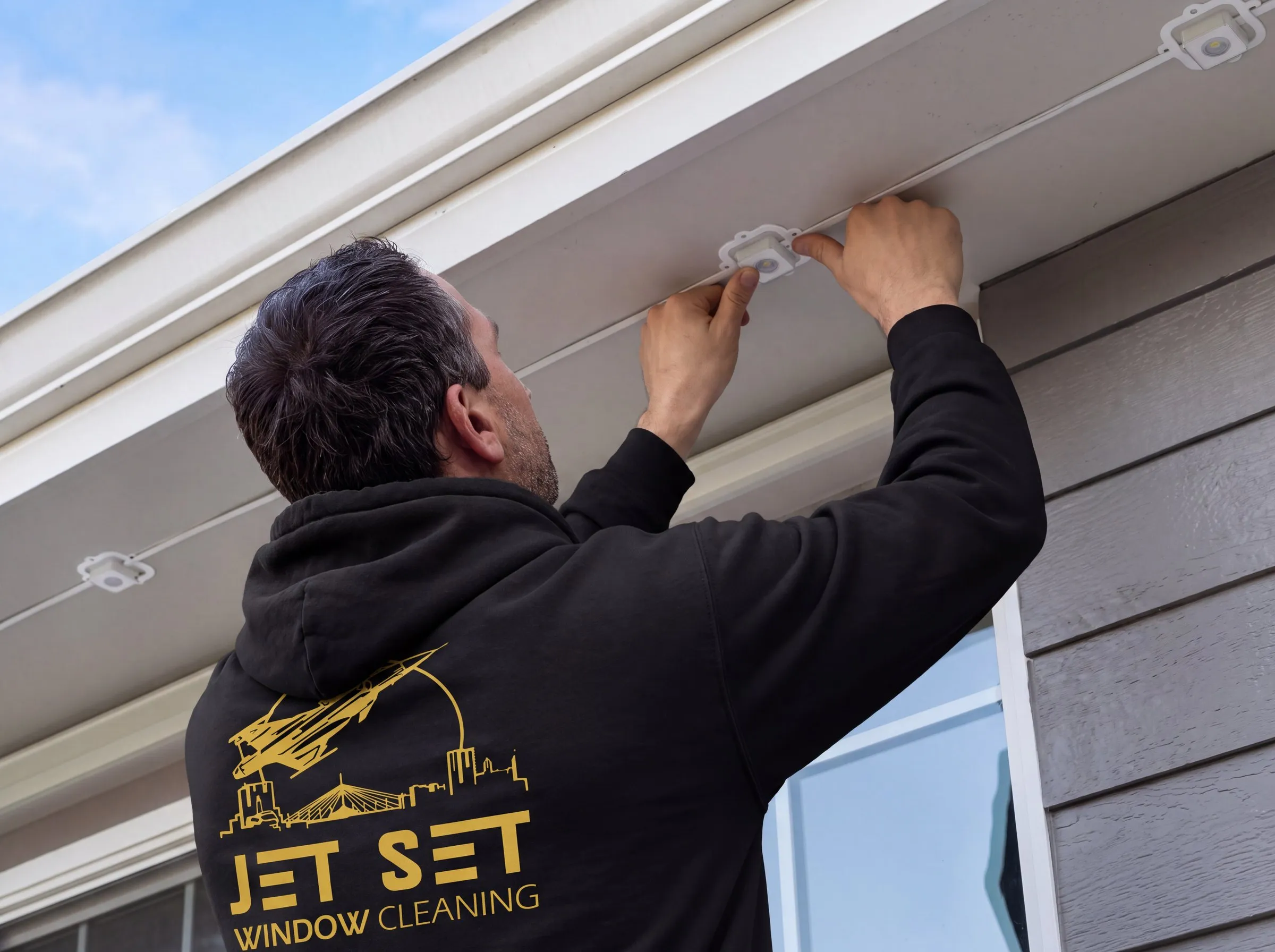 Jet Set installer in a black Jet Set hoodie pressing a Govee permanent light head onto the cable under the soffit of a Winnipeg home