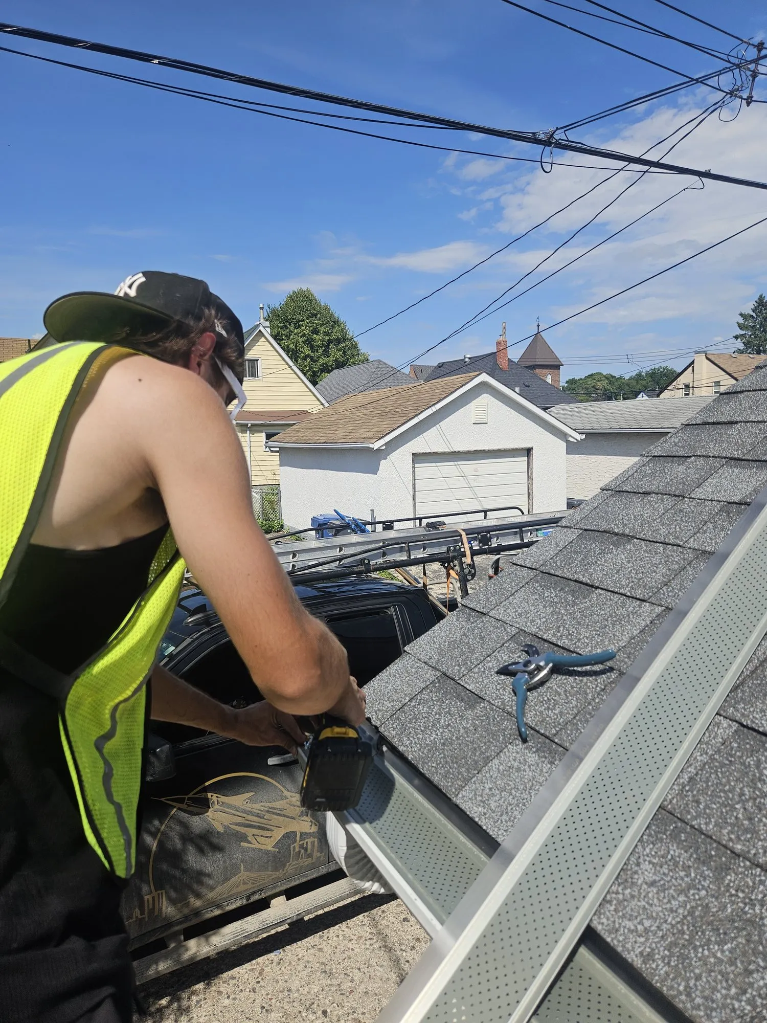 Jet Set technician in a hi-vis vest fastening a gutter guard to an eavestrough at the roof edge of a Winnipeg bungalow with a screw gun